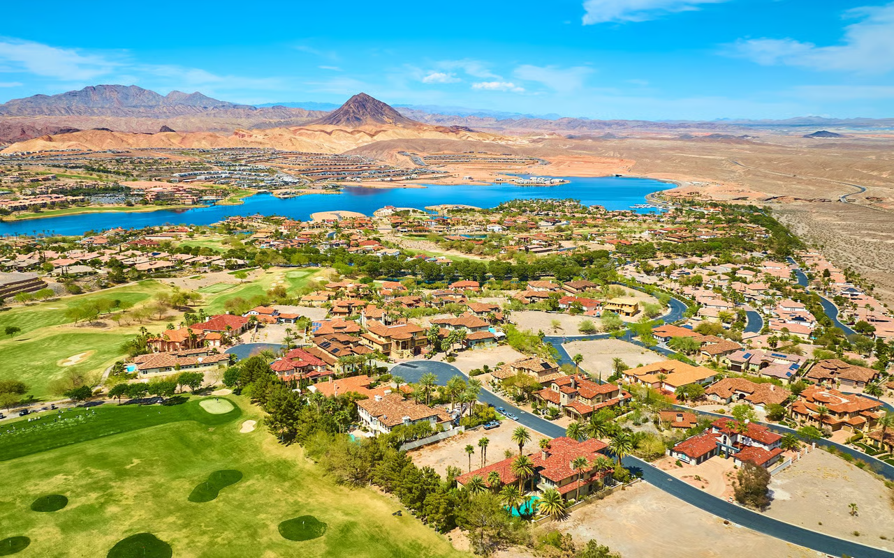 Aerial view of Skye Canyon in northwest Las Vegas with a blue lake, golf course, tile-roofed homes, and the desert mountain range beyond