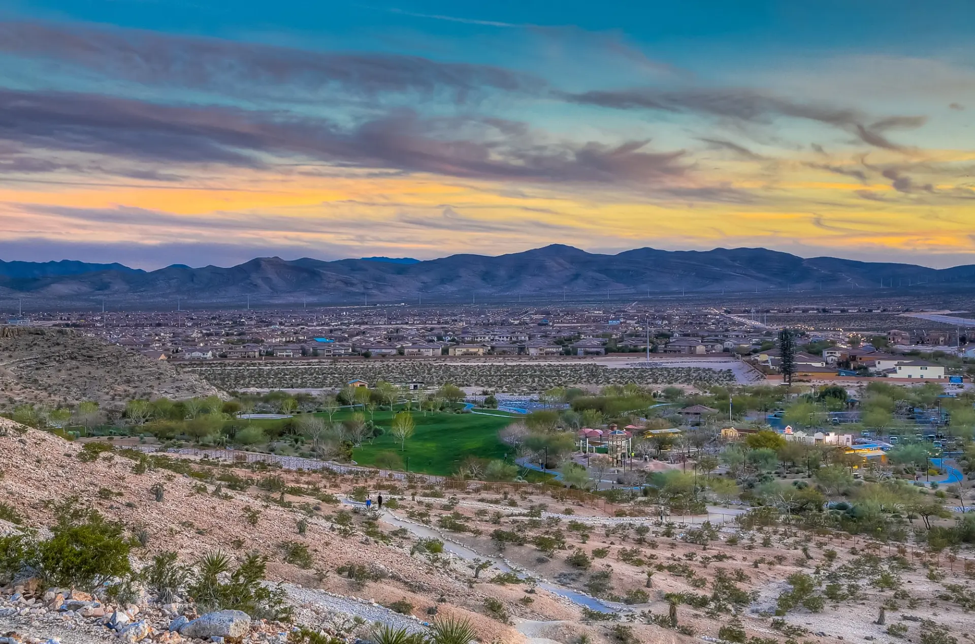 Aerial view of a Southwest Las Vegas community park with green lawns and trails, homes beyond, and desert mountains under a sunset sky